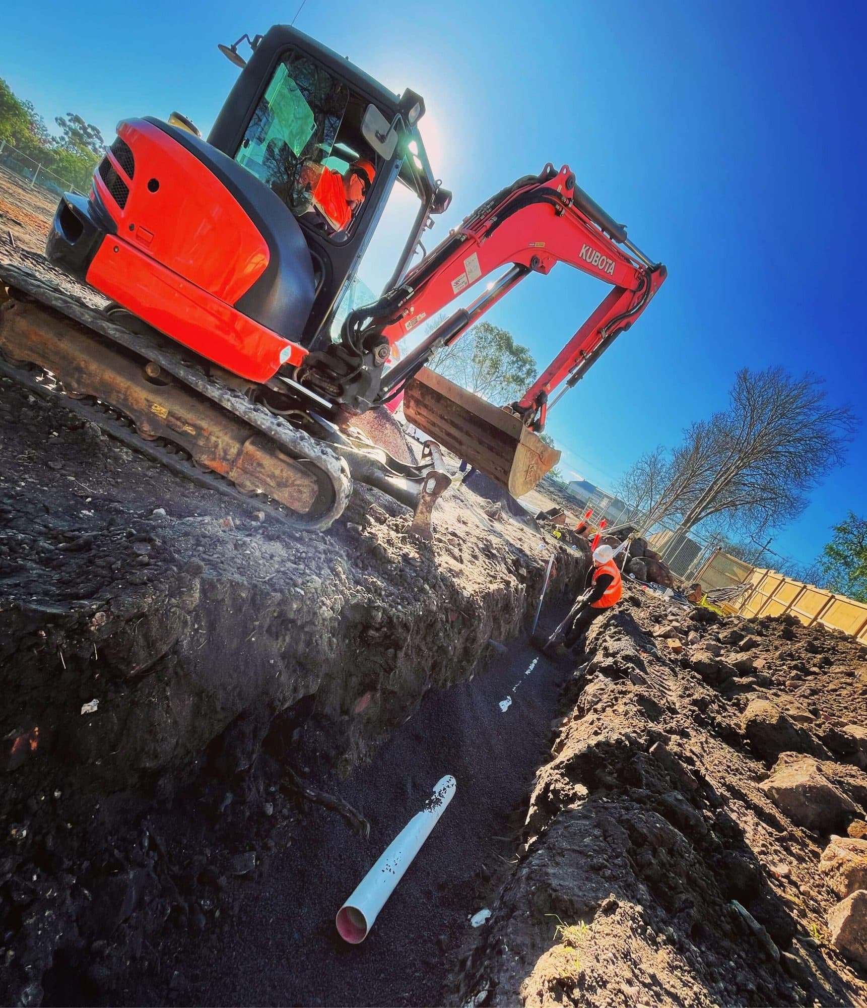 A red Kubota excavator digging a stormwater trench with fresh PVC pipe laid in